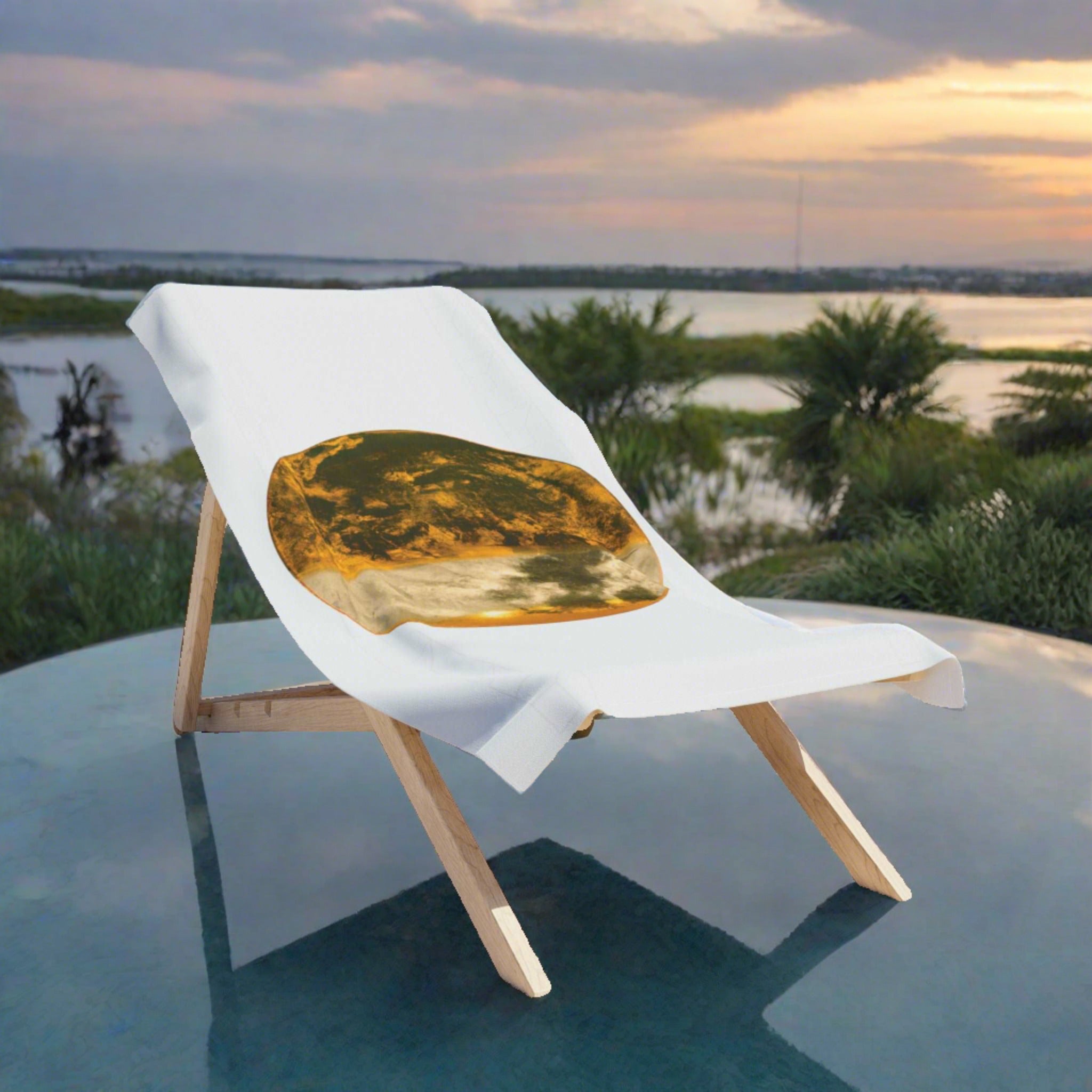 As evening falls, the Gilded Twilight Collection bath towel lies on a wooden deckchair standing on a round glass platform by a tranquil lakeside. The last rays of the sun cast a golden glow over the scene, mirroring the towel’s warm hues. The water and sky provide a serene backdrop of deepening blues and greens, highlighting the towel's rich design