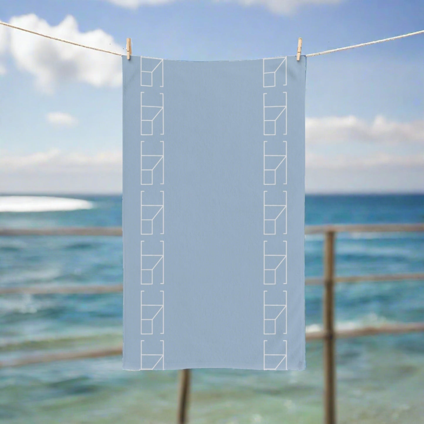 blue mist hand towel hanging from a rope outdoors, Imagine a relaxing beachside setting where the Blue Mist hand towel is hung by wooden clothespins on a rope strung between two weathered dock posts. The ocean's tranquil blues in the background mirror the towel's hue, and the gentle sea breeze animates the fabric, capturing the essence of a serene coastal retreat.