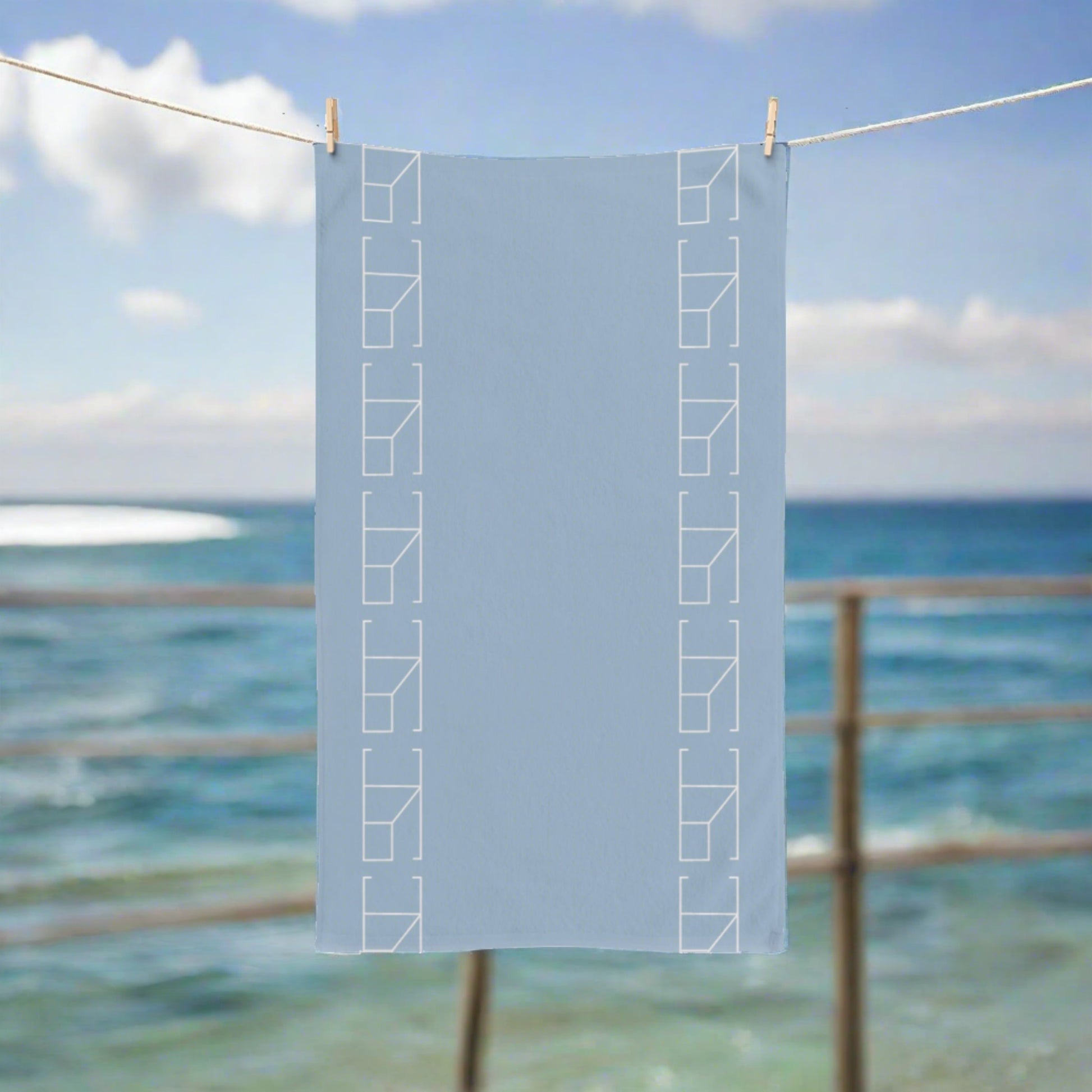 blue mist hand towel hanging from a rope outdoors, Imagine a relaxing beachside setting where the Blue Mist hand towel is hung by wooden clothespins on a rope strung between two weathered dock posts. The ocean's tranquil blues in the background mirror the towel's hue, and the gentle sea breeze animates the fabric, capturing the essence of a serene coastal retreat.