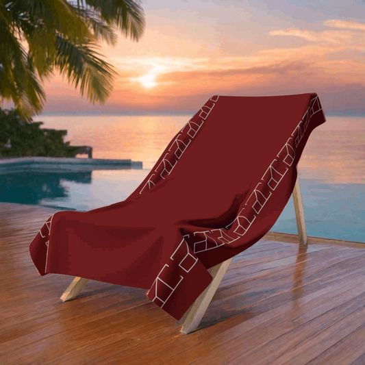 BarnRed28 Bath towel in a secluded and serene spa retreat background, where the red beach towel is draped over a bamboo lounge chair beside a tranquil infinity pool. The pool's water reflects the sky at sunset, offering hues of pink and orange that complement the red towel. Lush tropical foliage surrounds the area, and the flooring is made of polished dark wood, adding to the exclusive feel of the setting.